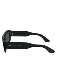 Black sunglasses with a sleek rectangular frame, plastic material, and sturdy arms. Features “Calvin Klein” branding and metallic accents.