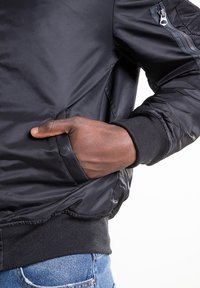 Zwarte bomberjas met een gladde textuur, geribbelde manchetten en een zak met rits op de bovenarm. Heeft een comfortabele pasvorm en een elastische zoom.