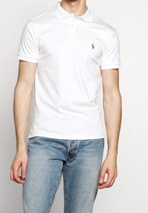 Mężczyzna w białej koszulce polo z krótkim rękawem i małym haftowanym logo, ubrany w jasnoniebieskie dżinsy na jednolitym tle.