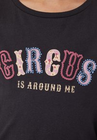 Zwarte shirt met kleurrijke decoratieve letters die "GIRLGS" spellen boven kleinere beige tekst "IS AROUND ME" op het borstgebied.
