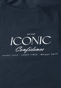 T-shirt en coton bleu marine avec un texte blanc : « CONFIANCE ICONIQUE » et « ÉTABLI EN 2025 », avec les détails « TEINTES DE COUCHER DE SOLEIL », « VIBRATIONS AMBRE », « JOURS ÉCLATANTS ». 
