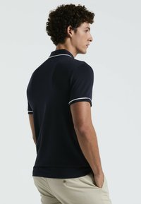 Polo shirt bleu marine avec des accents blancs, à manches courtes et col. Le tissu semble lisse. Le modèle présente le shirt de dos, les mains dans les poches.