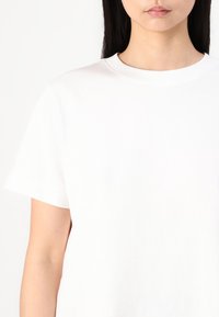 Jeune personne aux longs cheveux foncés portant un t-shirt blanc uni à manches courtes et col rond sur fond blanc.