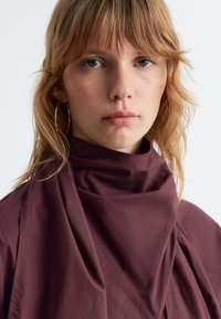 Dark burgundy high-neck top made of soft fabric, featuring draped texture and minimal seams for a clean, modern look.