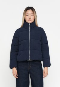 Kvinne iført en marineblå quiltet puffjakke med høy krage og mørk denimjeans, som står støtt mot en ensfarget hvit bakgrunn.