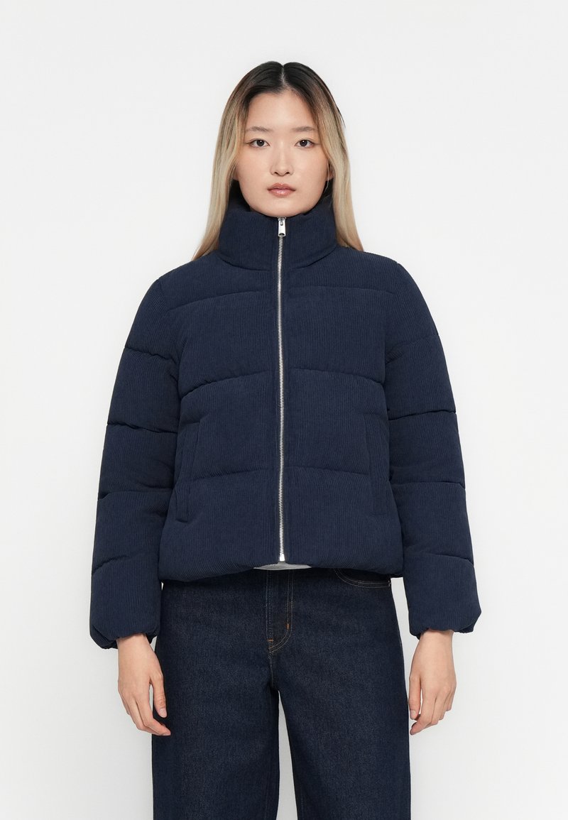 Mujer vestida con una chaqueta de plumas acolchada en azul marino con un cuello alto y jeans de mezclilla oscuros, de pie contra un fondo blanco liso.