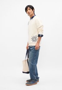 Ανοιχτό μπεζ φούτερ με ναυτικό τύπωμα ήλιου, τοποθετημένο πάνω από μπλε καρό πουκάμισο. Φοριέται με μπλε τζιν και γκρι αθλητικά παπούτσια, κρατώντας μία μπεζ τσάντα.