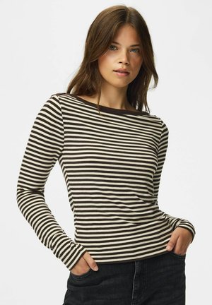 Jeune femme aux longs cheveux bruns portant un t-shirt à manches longues rayé noir et blanc et un jean noir, les mains dans les poches.