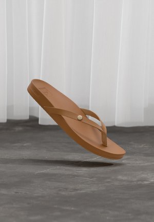 Brown flip-flop sandal with a button detail on the strap, floating above a gray concrete floor with white curtains in the background.