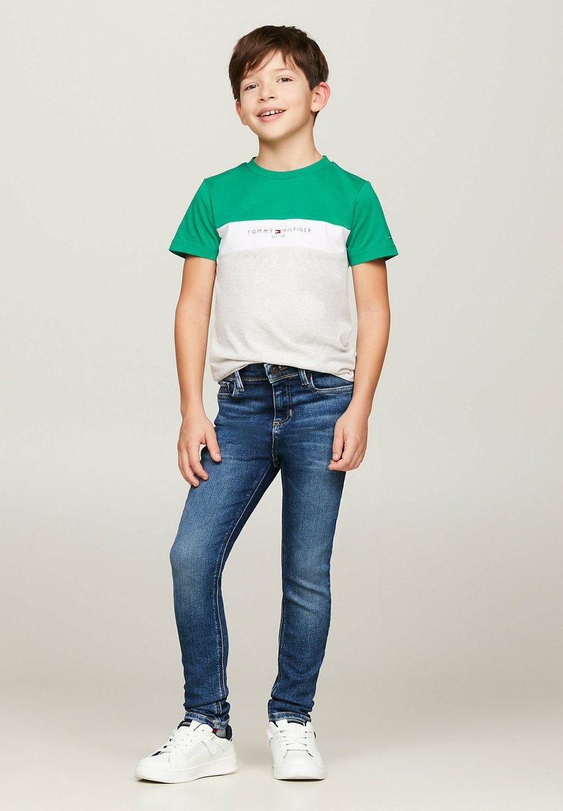 Zielona i szara koszulka z krótkim rękawem z logo, w połączeniu z dopasowanymi niebieskimi dżinsami i białymi sneakersami. Gładki materiał i swobodny styl.