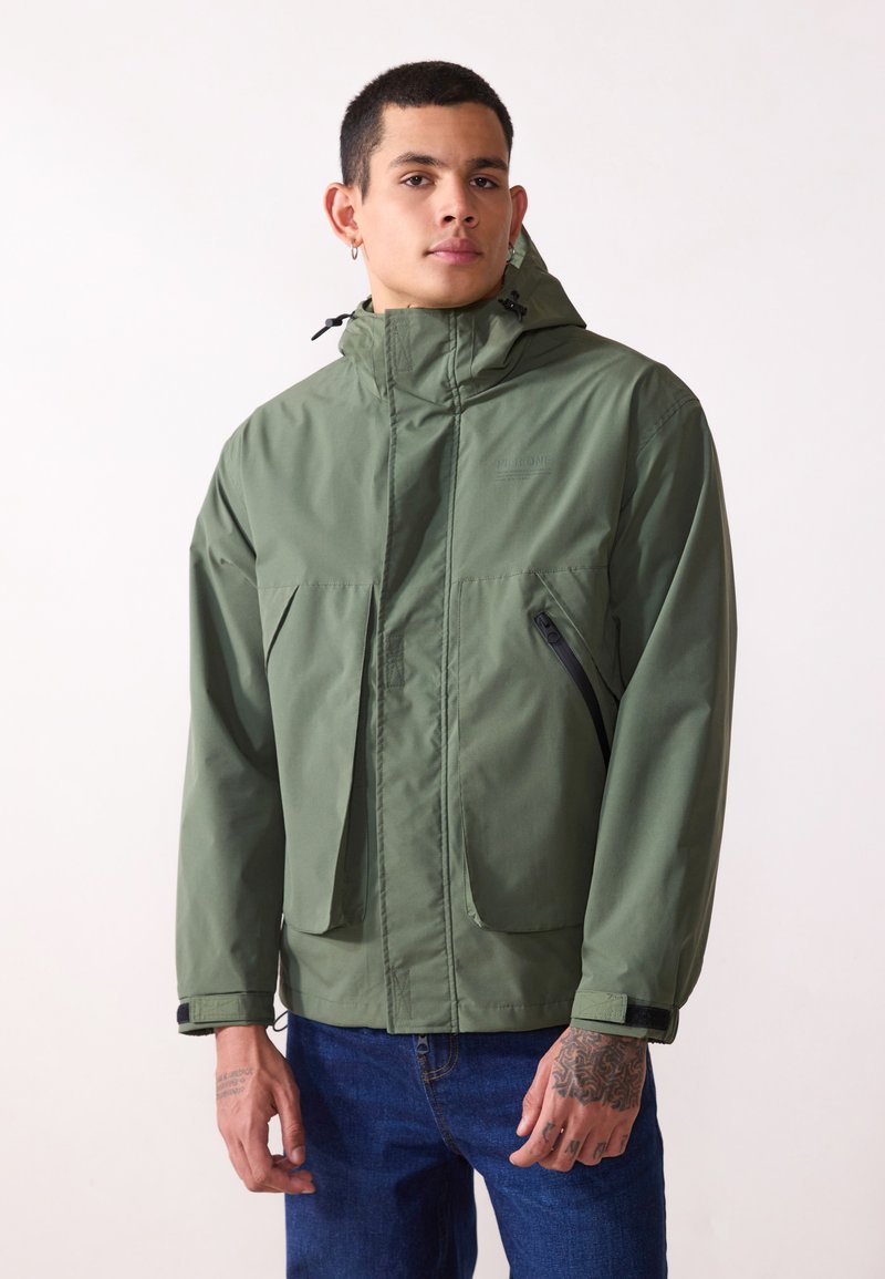 Veste imperméable verte avec un col haut, capuche réglable, poche latérale zippée et poignets élastiques. Texture lisse, design pratique.