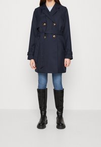 Trench coat azul marino con botones dorados de doble botonadura, cintura ceñida con cinturón, diseño ligeramente acampanado. Combinado con vaqueros azules y botas negras hasta la rodilla.