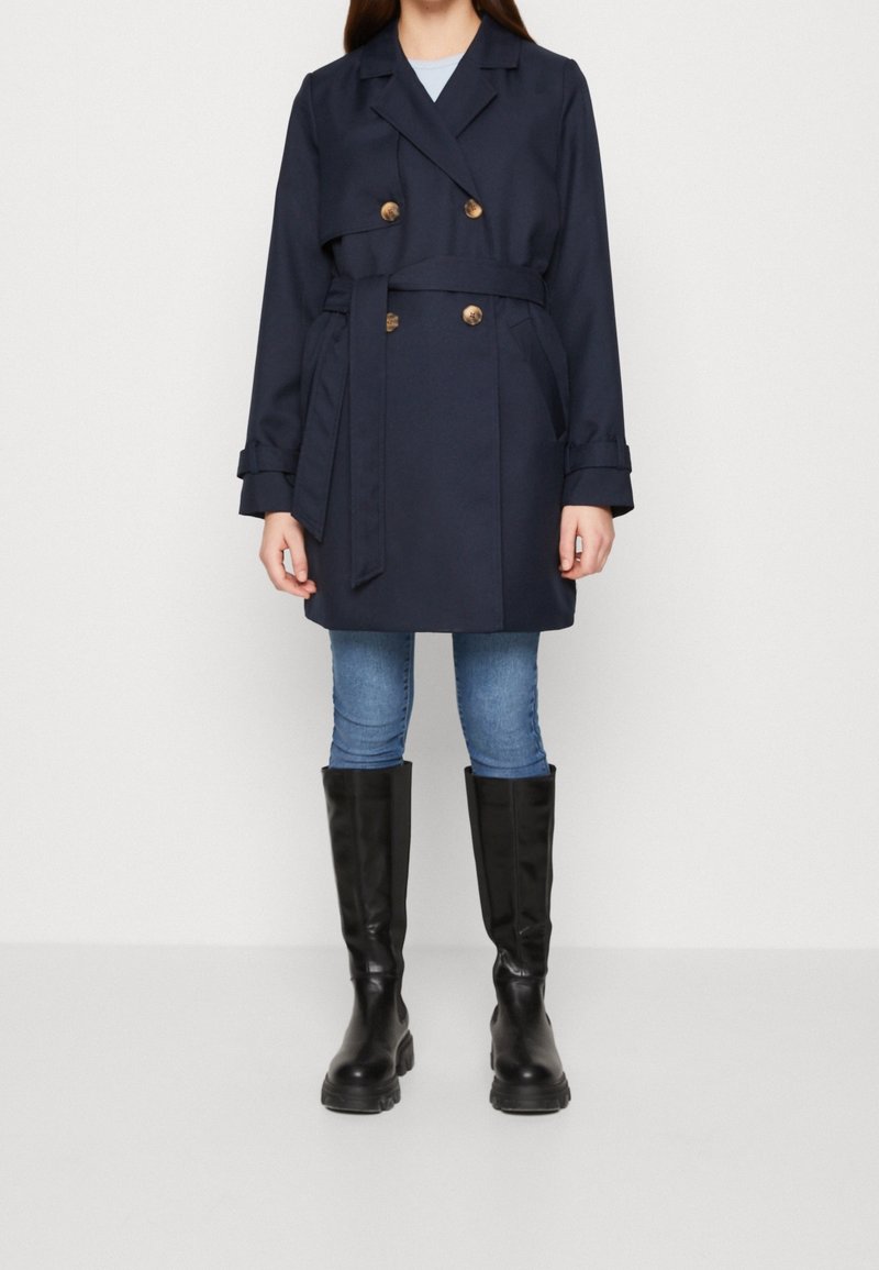 Trench coat azul marino con botones dorados de doble botonadura, cintura ceñida con cinturón, diseño ligeramente acampanado. Combinado con vaqueros azules y botas negras hasta la rodilla.