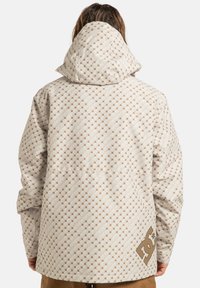 Osoba w wzorzystej beżowej kurtce z kapturem z logo DC na dolnej części pleców, stojąca tyłem na jednolitym tle.