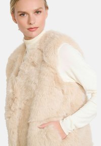 Gilet in pelliccia sintetica beige con una finitura morbida e testurizzata, dotato di tasche laterali e un design aperto, indossato sopra un dolcevita color crema.