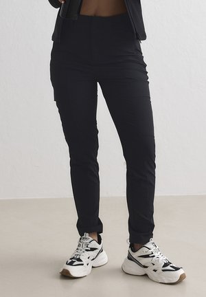 Person, die schwarze, eng anliegende Hosen und weiße und schwarze Sneaker trägt, steht auf einem hellen Boden vor einer weißen Wand.