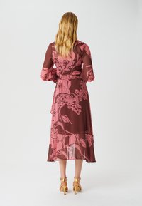 Larga y etérea vestido floral en rosa intenso y burdeos, con un cinturón en la cintura y mangas largas. Complementado con sandalias de tacón doradas.