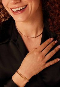 Roségouden en roze kralenketting gelaagd met een gedraaide metalen ketting, gedragen met een gouden en roze armband op een donkere top.