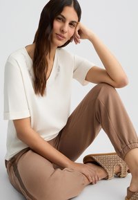 Haut crème à col en V avec des manches courtes, présentant des accents brodés. Pantalons marron fuselés avec des poignets élastiques. Sandales à motifs beige.