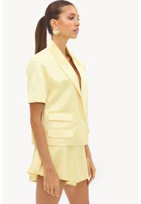 Femme avec queue de cheval brune portant de grandes boucles d'oreilles dorées à motif floral, un blazer jaune pâle à manches courtes et une jupe plissée assortie, tournée vers la droite.