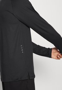 Osoba v černém dlouhém rukávu značky Puma se natahuje s rukama do stran, přičemž ukazuje detail bočního švu a logo Puma na levé straně.