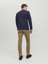 Blazer bleu marine avec une coupe ajustée, chinos beige et chaussures noires. Le blazer a une texture lisse et des revers classiques, avec des détails de couture visibles.