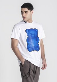 Maglietta bianca a maniche corte con una grande grafica di un orsetto di gelatina blu lucido sul davanti. Il modello indossa pantaloni larghi di colore chiaro.