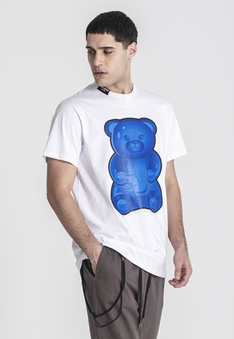 Maglietta bianca a maniche corte con una grande grafica di un orsetto di gelatina blu lucido sul davanti. Il modello indossa pantaloni larghi di colore chiaro.
