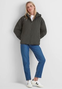 Woman wearing a dark quilted hooded jacket, blue jeans, and white sneakers, standing against a plain light background with hands behind back.