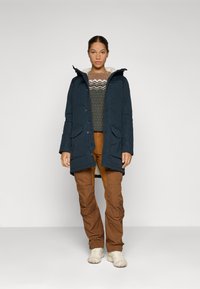 Marinemarineblauwe parka-jack met capuchon en klepzakken, gedragen over een gestreept shirt en een bruine broek, aangevuld met lichtgekleurde schoenen.