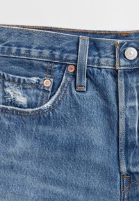 Närbild av midjan på blå denimjeans med bältesögle, metallknapp, nitar och en liten sliten framficka med fransat tyg.