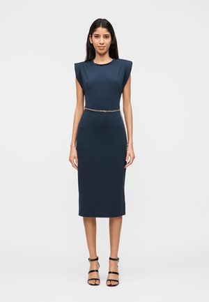 Moteris dėvi prigludusią tamsiai mėlyną suknelę be rankovių, su paminkšlintais pečiais ir aukso spalvos grandinėlės diržu, derinama su juodais sandalais su dirželiais.