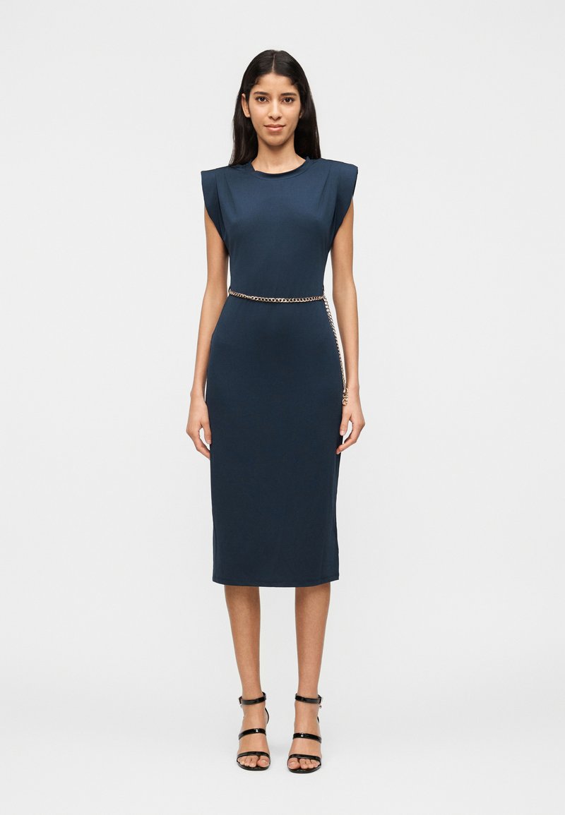 Γυναίκα που φοράει στενή αμάνικη μπλε φόρεμα με μαξιλαρωμένους ώμους και χρυσή αλυσίδα στη μέση, συνδυασμένη με μαύρα πέδιλα με λουριά.