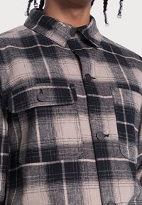 Kostkovaná flanelová košile s béžovo-černým barevným schématem, dvěma kapsami na prsou, knoflíkovým zapínáním a standardním límečkem.