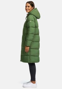 Green puffer coat with a hood, featuring quilted design and side pockets. Worn over black jeans and white sneakers.