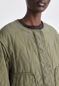 Pikowana oliwkowa kurtka w diamentowy wzór, z okrągłym dekoltem i dwiema kieszeniami. Miękka tekstura i stonowany design.