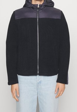 Osoba w czarnej polarowej kurtce z ciemnymi pikowanymi ramionami i kapturem, zapinana z przodu na zamek, zestawiona z jasnoniebieskimi jeansami.