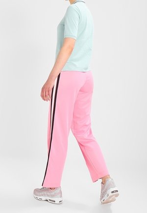 Osoba w różowych spodniach dresowych z czarnymi bocznymi pasami, jasnoniebieskiej koszulce z krótkim rękawem i beżowych trampkach, stojąca bokiem na białym tle.