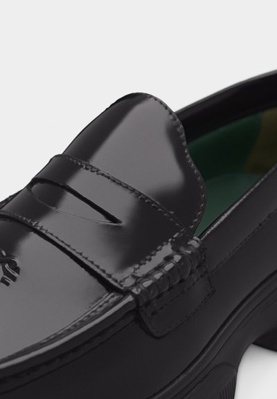 Γυαλιστερό μαύρο δερμάτινο μοκασίνι με ραμμένες λεπτομέρειες και λουράκι τύπου penny πάνω από το επάνω μέρος, εμφανιζόμενο σε κοντινή λήψη.