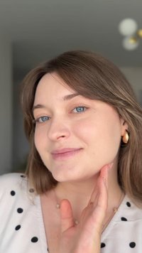 Nærbilde av en person med lys hud, blå øyne, langt brunt hår, iført gull hoops øredobber og en hvit skjorte med svarte prikker.