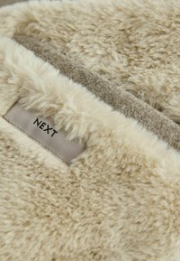 Beige kunstpelsstof med en blød tekstur, en glat brun bagside og et syet mærke med teksten "NEXT."