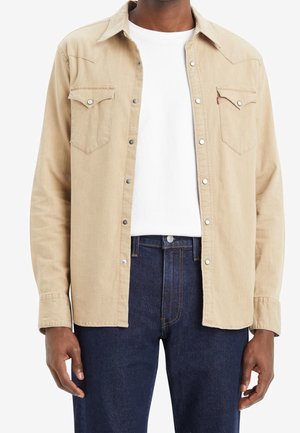Man met een beige overhemd met knopen over een wit T-shirt en donkerblauwe spijkerbroek, staand met ontspannen armen langs zijn zij.