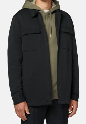 Mann trägt eine schwarze Jacke mit Knöpfen über einem olivgrünen Kapuzenpullover mit Kordelzug, steht mit entspannt an den Seiten hängenden Händen.