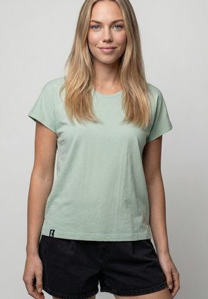 Frau mit langen blonden Haaren, die ein hellgrünes kurzärmliges Hemd und schwarze Shorts trägt, vor einem schlichten grauen Hintergrund stehend.