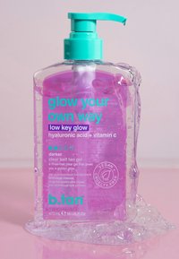 Bouteille de pompe transparente contenant un gel violet étiqueté "brillez à votre manière" auto-bronzant avec acide hyaluronique, vitamine C, végan et sans cruauté, sur une surface rose.