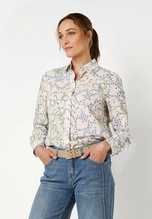 Frau mit braunen Haaren im Pferdeschwanz, die ein geblümtes Hemd mit Knöpfen, einen beigen Gürtel und blaue Jeans trägt, mit den Händen in den Taschen stehend.