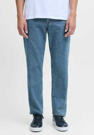 Homme portant un jean bleu droit, un t-shirt blanc et des baskets noires à semelles blanches, debout devant un fond clair uni.