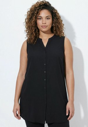 Kobieta o kręconych włosach nosi bezrękawną czarną tunikę z guzikami, stoi na białym tle.