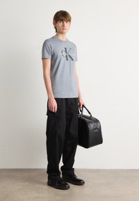 Gray t-shirt with "Calvin Klein Jeans" logo, black cargo pants, black shoes, and a black leather bag with curved edges and a handle.