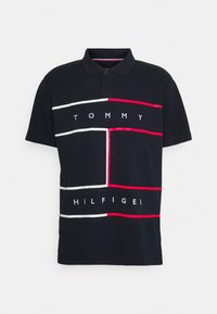 Musta polo-paita, joka on valmistettu puuvillasta, punaisilla ja valkoisilla geometrisilla raidoilla, ja tekstillä "TOMMY HILFIGER" lihavoidulla valkoisella fontilla.